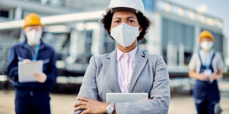 African American building contractor with protective face mask standing with her arms crossed and looking at the camera. Two civil engineers are in the background.